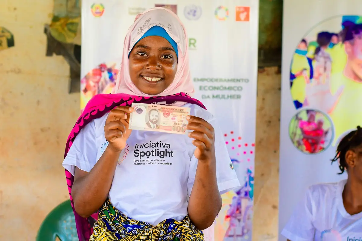 Jovem mulher com camisola da Iniciativa Spotlight mostra uma nota, diante de uma faixa sobre empoderamento económico da mulher